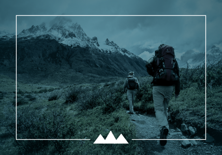 Two hikers on a mountain trail, one leading the way toward snow-capped peaks
